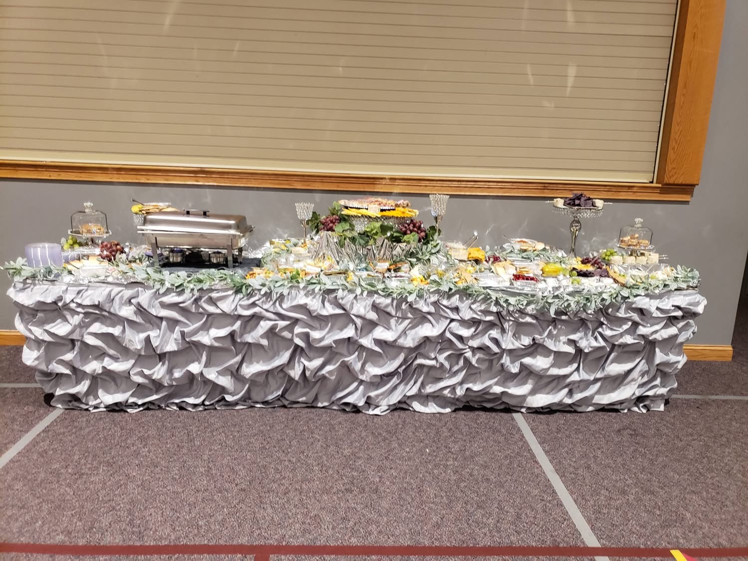 Catered buffet table dressed in silver ruffled linen with greenery and fresh hors d'oeuvres